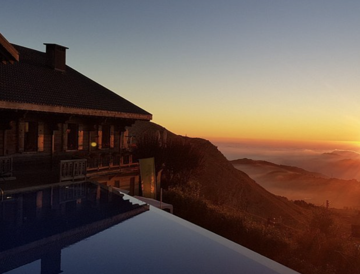 A house with a tiled roof on a mountain at sunset, overlooking a valley with fog and distant mountains.
