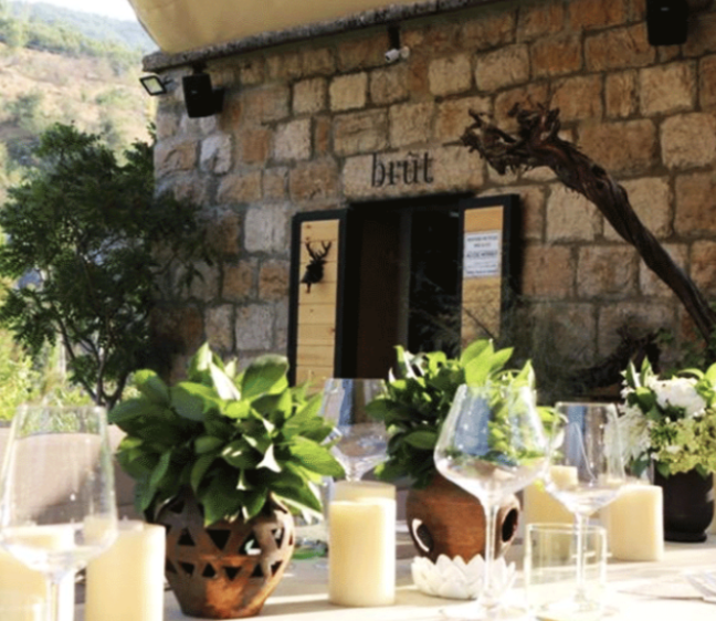 Decorative table setting with glassware, candles, and potted plants outside a rustic stone building with a door labeled 'brut' in the background.