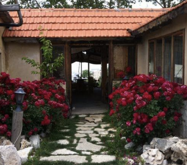 Pathway leading to a rustic house entrance, flanked by pink and red flowering bushes, with a small lamp post on the left.
