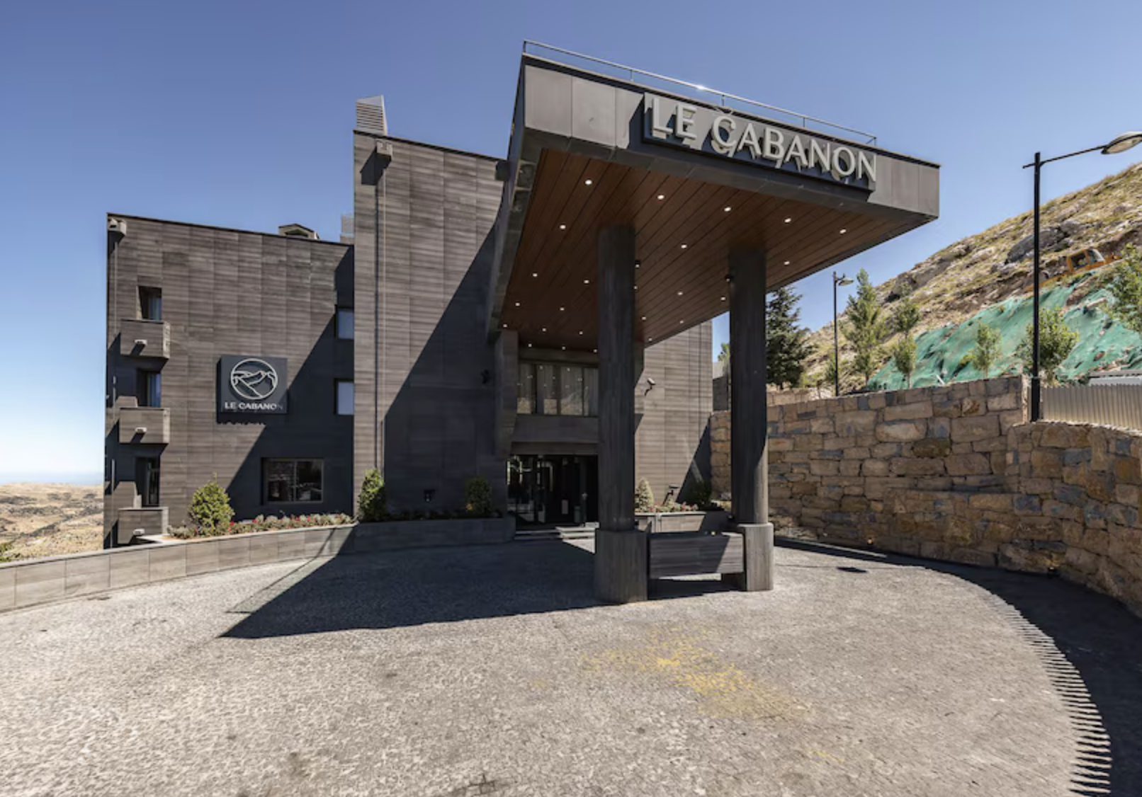 The entrance of Le Cabanon hotel, a modern building with dark exterior, large sign with the hotel's name, an overhang supported by wooden columns, and a small landscaped area with plants and flowers.