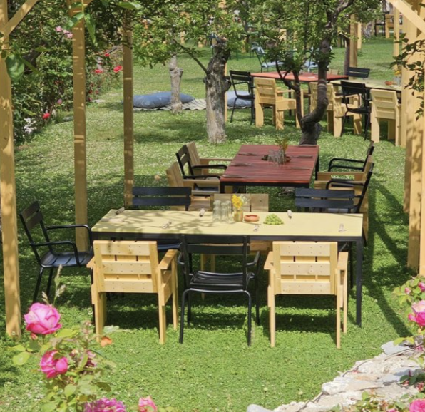 Outdoor garden with wooden and black metal tables and chairs, surrounded by trees and pink flowers.