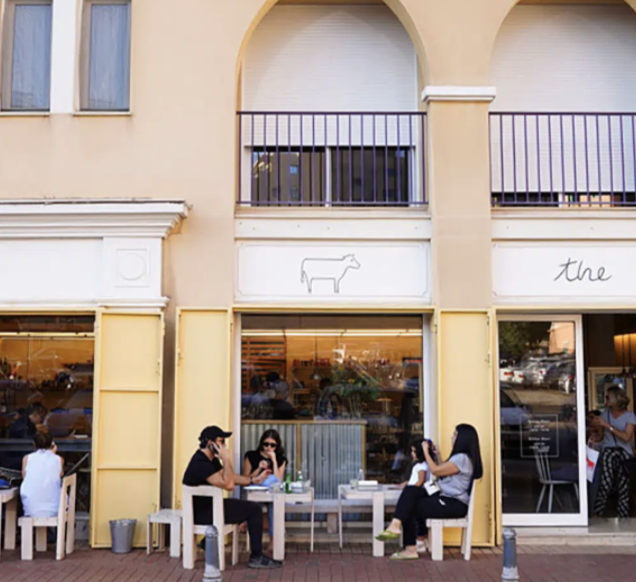 People sitting outside a cafe with large windows and a storefront sign featuring a cow outline and partial text 'the' on a yellow building.