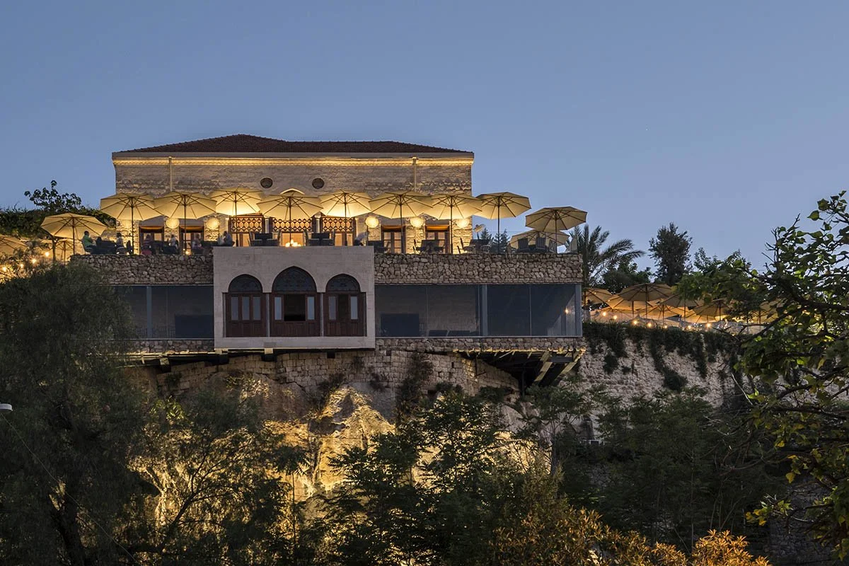 A building on a hill at dusk, illuminated with warm lighting, has outdoor seating with umbrellas and a glass wall, surrounded by trees.