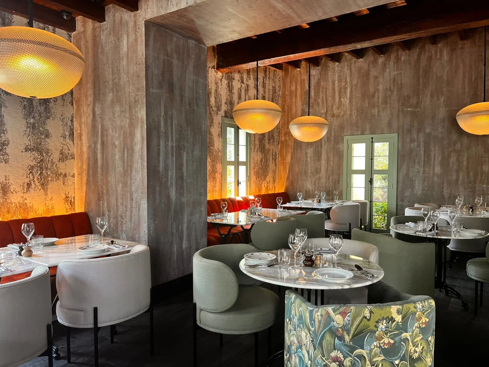 A cozy restaurant interior with round tables set for dinner, with white plates, glasses, and cutlery. The room has textured gray walls, wooden beams on the ceiling, and modern hanging pendant lights. There are windows letting in natural light, and a mix of upholstered chairs and booths in neutral and muted tones.