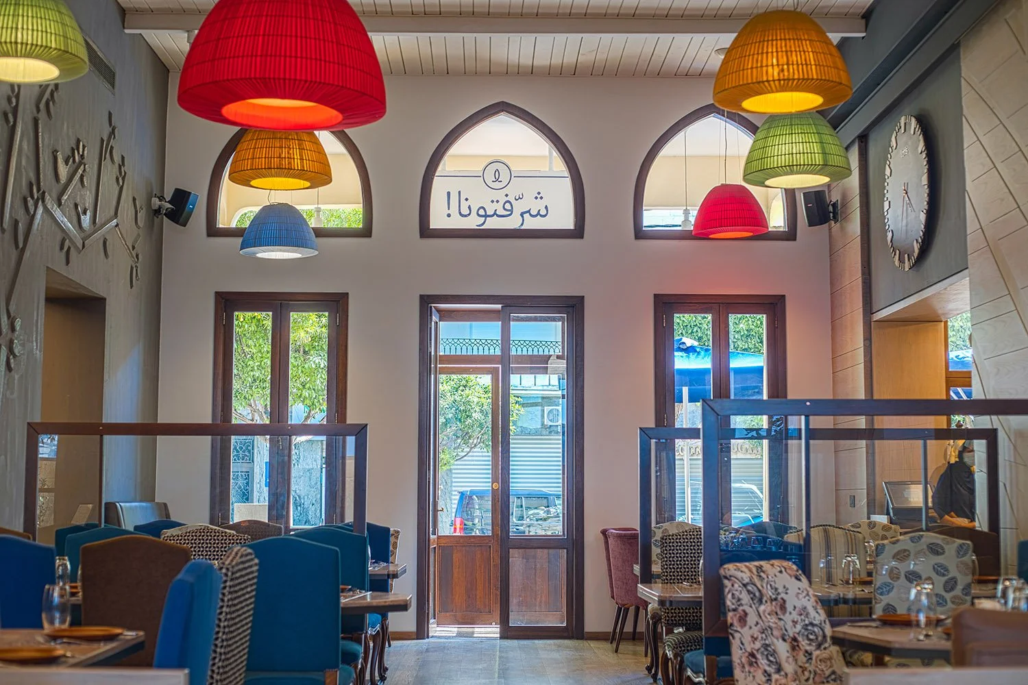 Interior of a restaurant with colorful hanging lanterns, wooden and patterned chairs, and large windows with a door in the center. A sign with Arabic script and a logo is displayed above the windows.