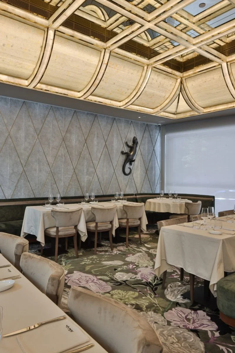 Interior of an upscale restaurant with boxed ceiling lighting, a large wall-mounted sculpture of a lizard, and elegant dining tables set with glassware, plates, and silverware on a floral-patterned carpet.