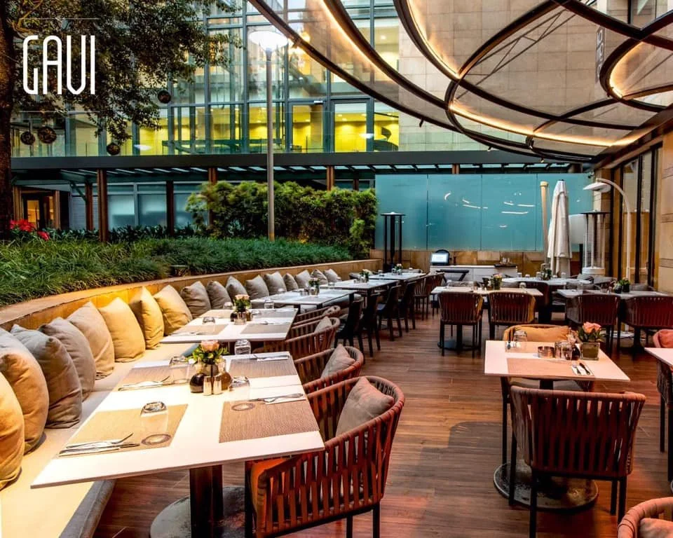 Empty outdoor dining patio with tables, chairs, and a cushioned bench, decorated with small flower arrangements, during evening setting with modern architecture and overhead lighting.