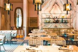 A stylish restaurant interior with wooden walls, open shelving displaying glassware, modern pendant lighting, and a large window with a curtain. The space features wicker chairs and a dining table.