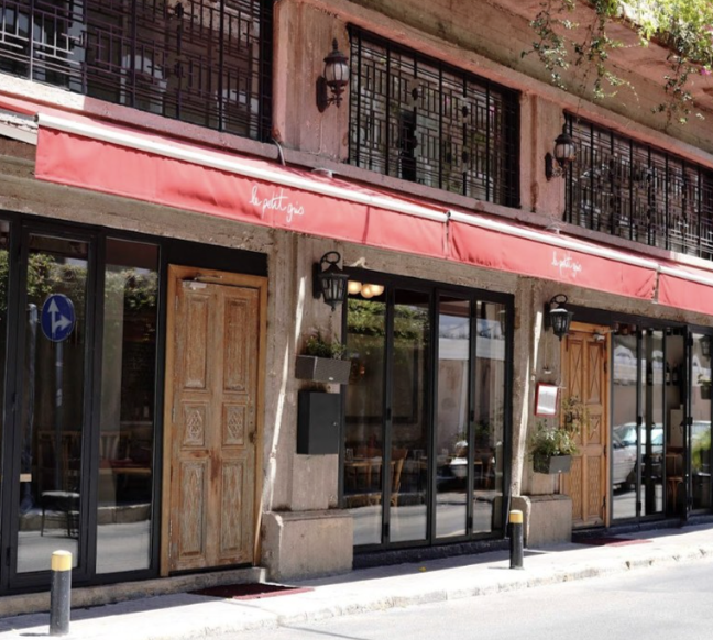 The exterior of a restaurant with glass windows, wooden doors, a red awning, and potted plants outside.