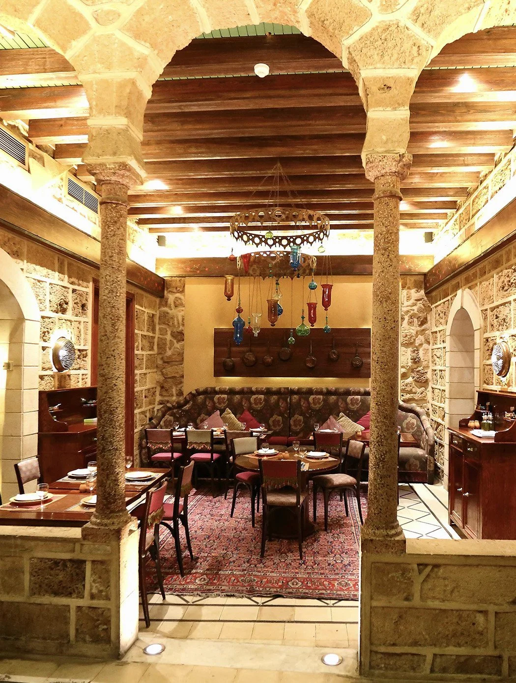 Interior view of a cozy restaurant with stone walls, wooden ceiling beams, and a chandelier made of hanging colorful glass bottles. There are wooden tables and chairs with pink and beige cushions, a patterned rug, and decorative plates on the walls.