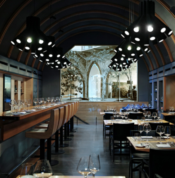 Elegant restaurant dining area with modern lighting, set tables with wine glasses, and a large window with an outdoor view of trees and a historic building.