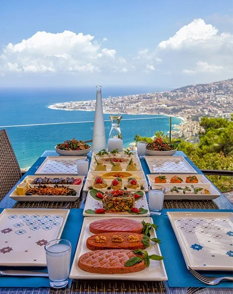 Elegant outdoor table set with various dishes and drinks, overlooking a scenic coastal view with the ocean and cityscape in the distance under a partly cloudy sky.