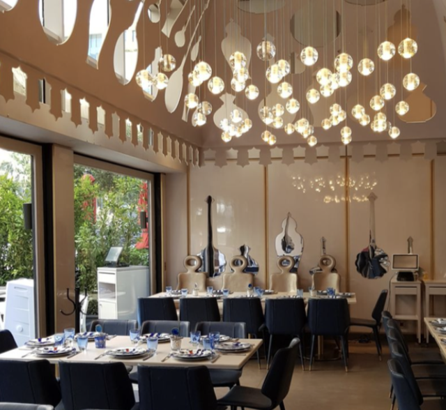 Elegant restaurant interior with modern hanging light fixtures, decorated tables with plates and glasses, and a large window showing greenery outside.
