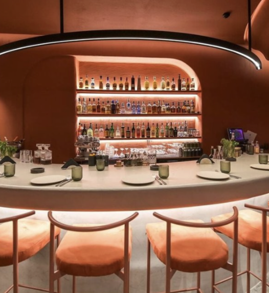 A modern bar with a curved counter, four orange chairs, and shelves filled with various bottles of alcohol backlit with warm lighting.