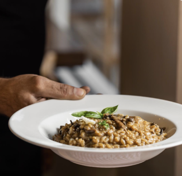 A person holding a white bowl filled with mushroom risotto, garnished with herbs.