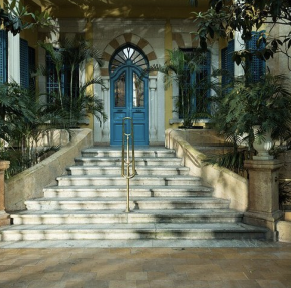 Marble staircase leading up to a bright blue door with an arched window above, flanked by lush palm plants on either side, outside a building with classical architecture.