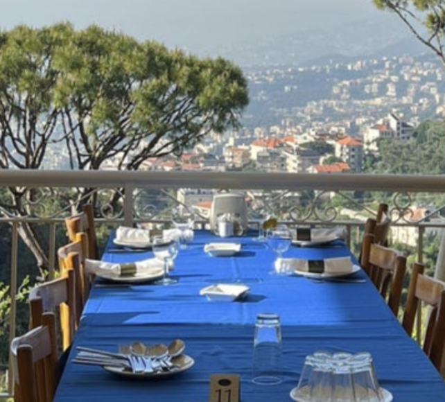 An outdoor dining table set for a meal with chairs around it, on a balcony with a view of a city and trees in the background.