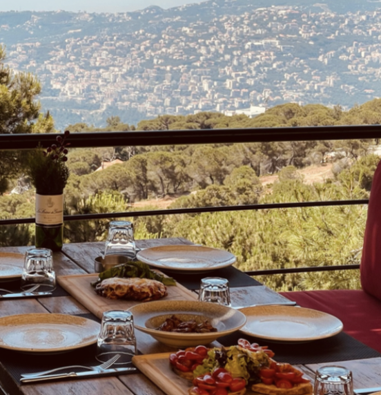 Outdoor dining table with plates, glasses, and food, overlooking a scenic landscape with hills and trees.