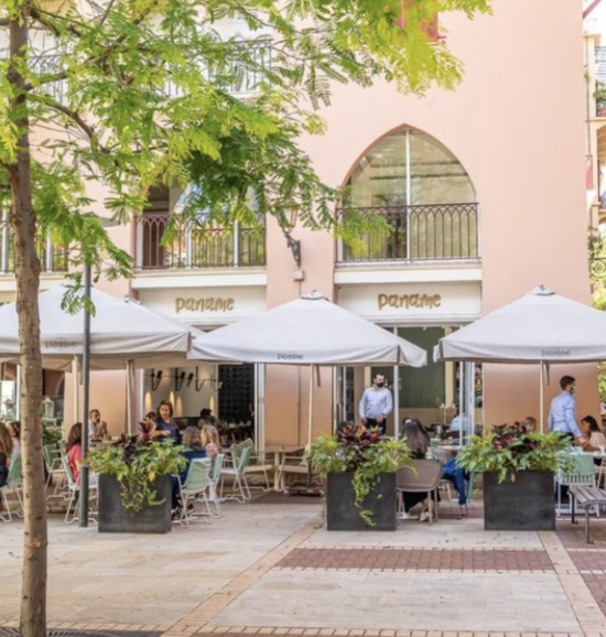 People dining outside at a restaurant with large umbrellas and potted plants on a city sidewalk, with a pink building in the background.