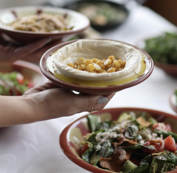Person holding a small bowl of hummus topped with chickpeas and olive oil at a table with other salads and dishes.