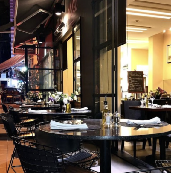 Outdoor patio seating at a restaurant with tables set with plates, napkins, and condiments, decorated with flowers, and an indoor dining area visible through large glass windows.