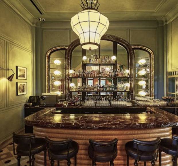 Elegant bar with a marble countertop, dark wood bar stools, and a backlit shelving unit filled with alcohol bottles, in a vintage-style room with framed pictures and green-paneled walls.