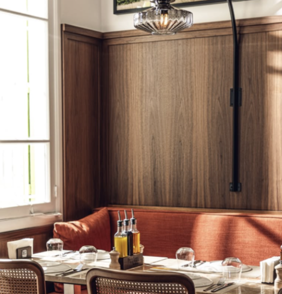 Dining table set with glasses, bottles of olive oil, and place settings, next to a red cushioned bench and a wood-paneled wall in a restaurant interior.