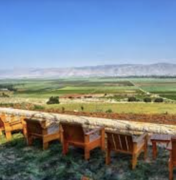 A scenic view of a landscape with rolling hills and fields under a clear blue sky, seen from a wooden terrace with chairs.