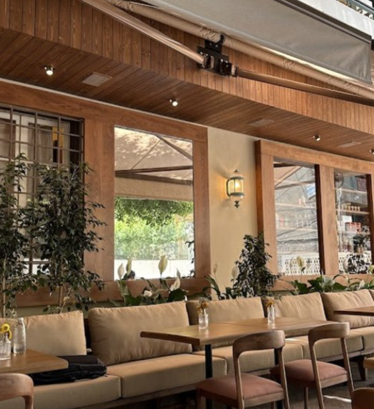 Interior view of a cozy restaurant or cafe with wooden accents, large windows, lush green plants, beige cushioned seating, and wooden tables with chairs.