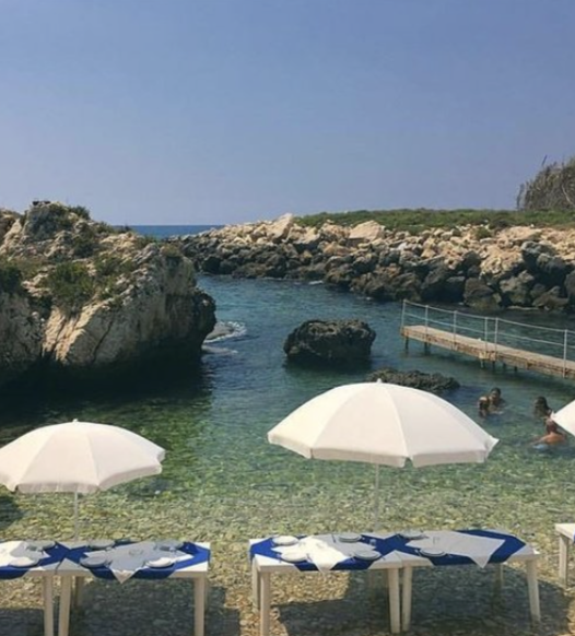 Beach scene with white umbrellas, lounge chairs, rocky shoreline, clear blue water, and people swimming.