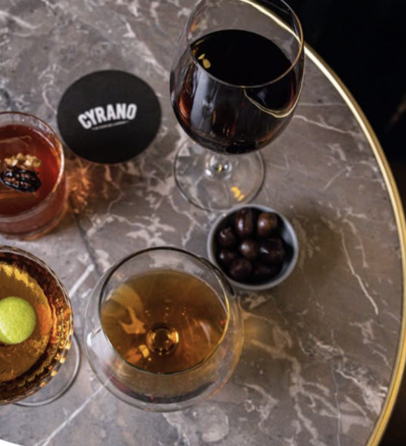 Three glasses of alcohol, two with dark red or brown liquids and one with a golden liquid, a bowl of round chocolates, and an informational card with "CYRANO" written on it, all on a marble table.