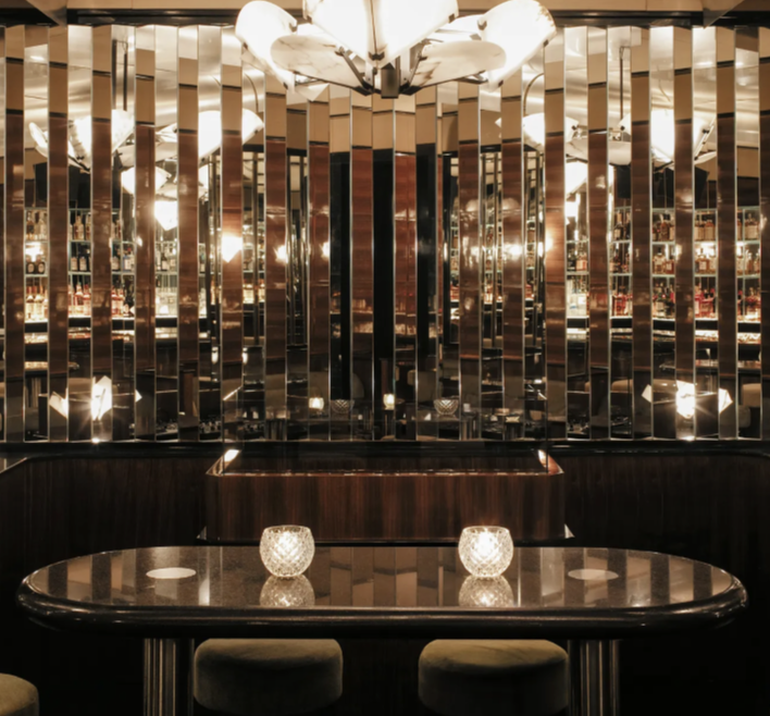 A luxurious, dimly lit restaurant or lounge with a reflective mirror wall, a dark polished table with two decorative candles, and a vintage chandelier overhead.