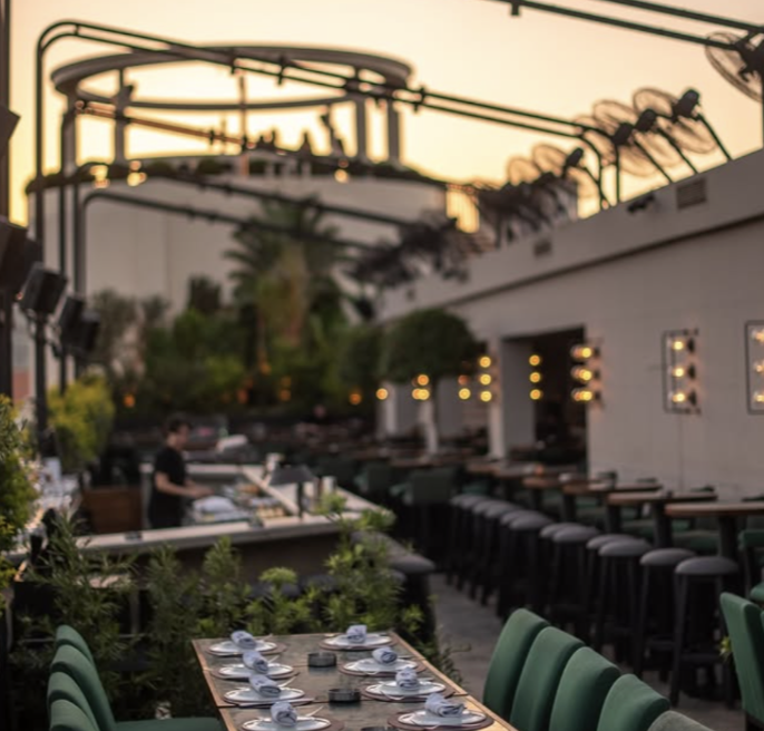 Outdoor restaurant patio at sunset with a long table set with plates, napkins, and glasses, green chairs, bar stools along the counter, string lights, and a person preparing food at the bar.