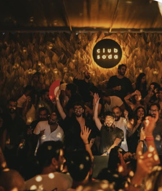 Crowd of people enjoying a party at Club Soda, with warm lighting and a textured wall backdrop. Some individuals have their hands raised, smiling and dancing.