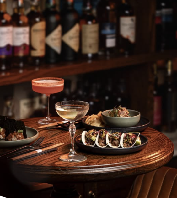 Two cocktails and a plate of tacos on a wooden table in a bar setting.
