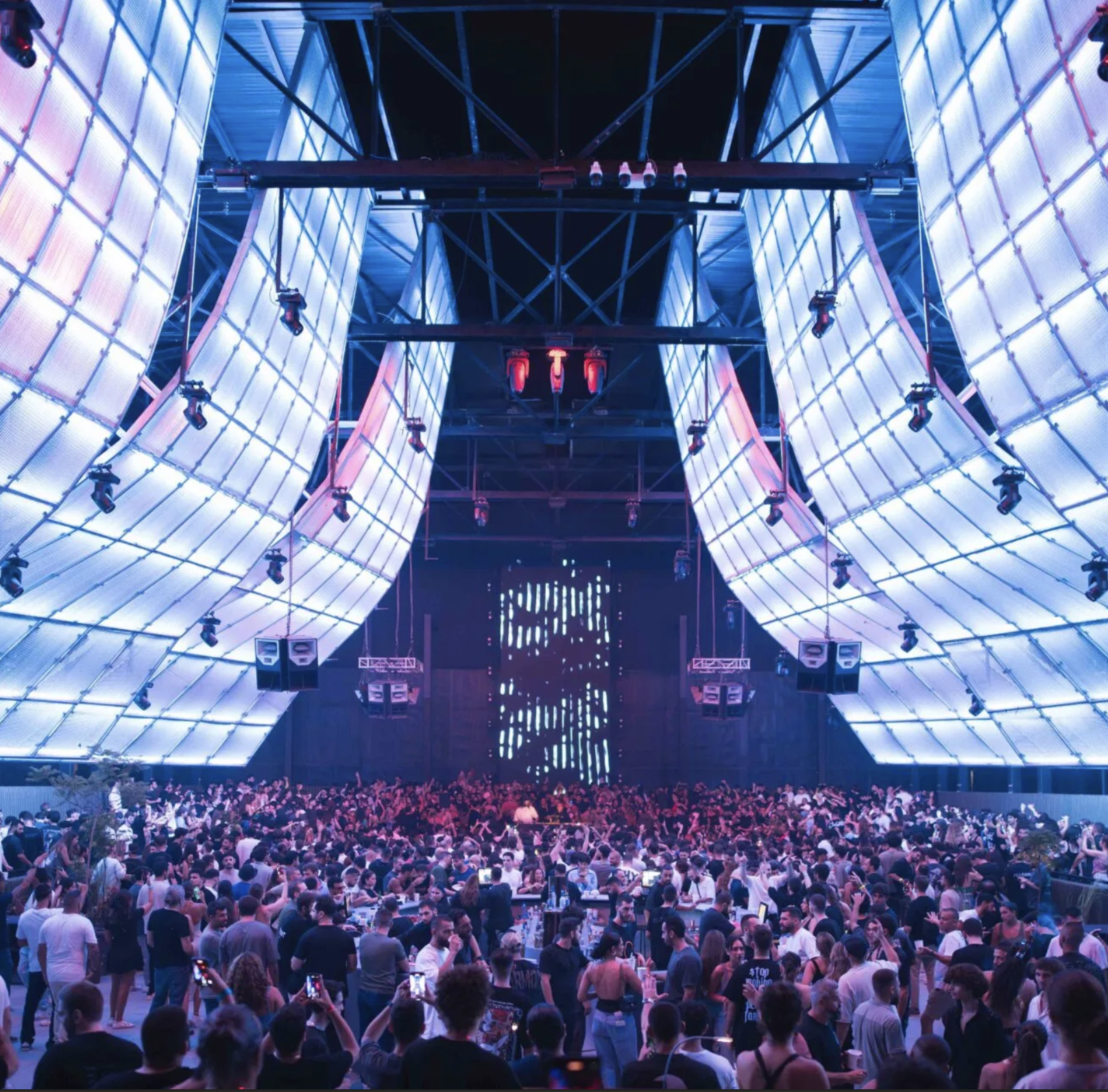 Large crowd of people at a concert or nightclub with blue and purple lighting, a stage with lighting effects, and a modern, indoor venue with high ceilings.