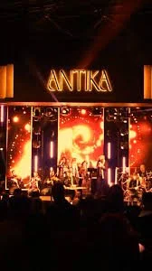 Concert stage with large LED screens displaying fiery visuals, illuminated 'ANTIKA' sign overhead, audience in foreground.
