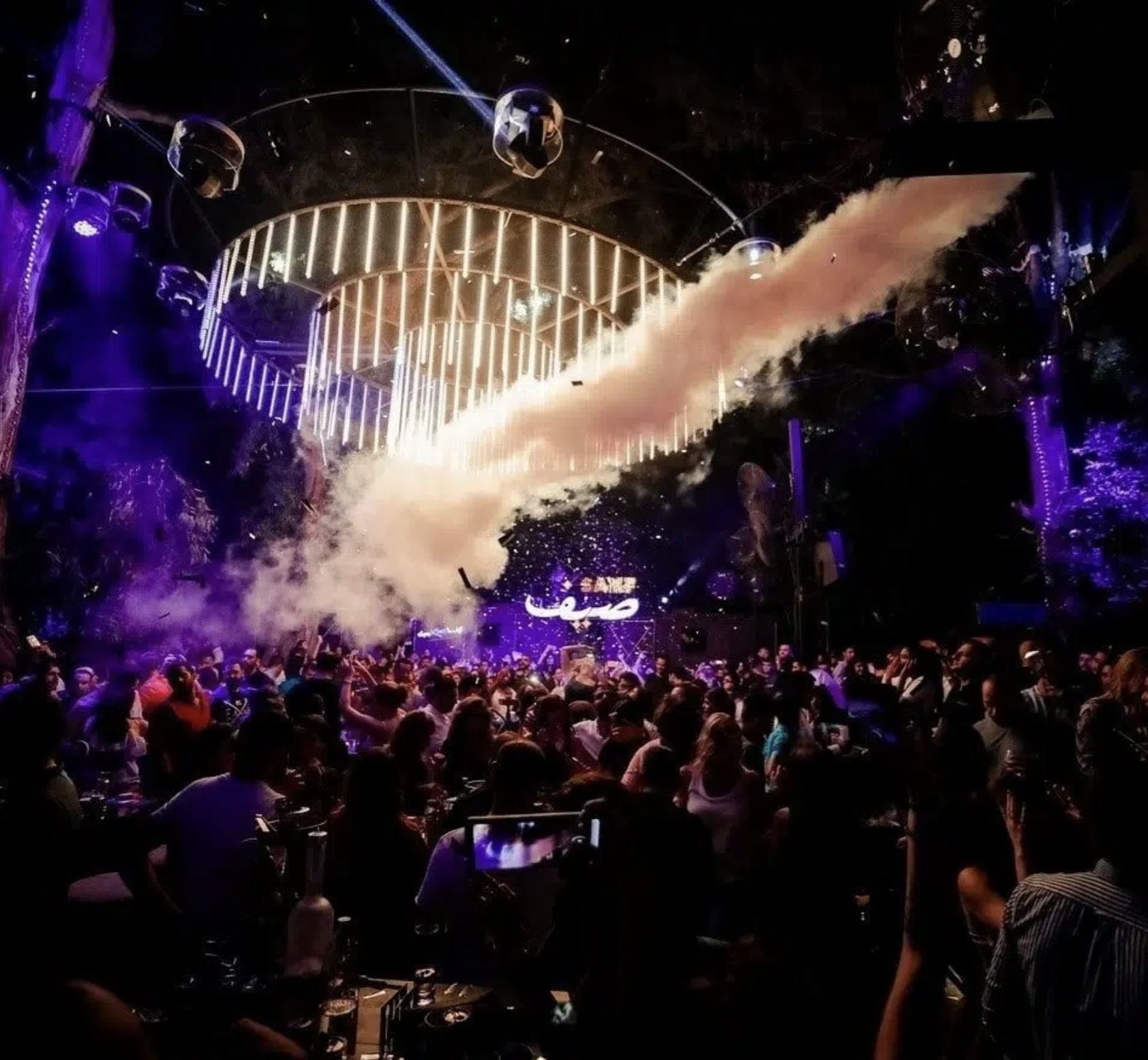 Crowd of people at a nightclub with a stage, neon lights, smoke effects, and a DJ booth.