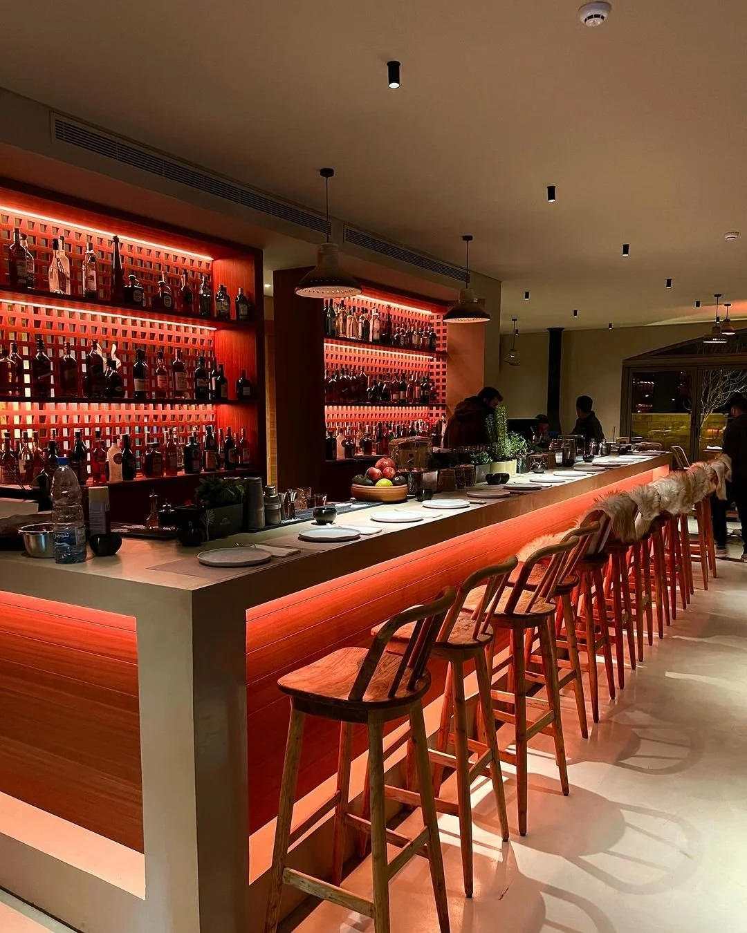A modern bar with a wooden counter, red accent lighting, and a backlit shelf filled with various bottles of alcohol. Several wooden bar stools with fur-covered backs are lined up along the counter, and patrons are seen in the background.