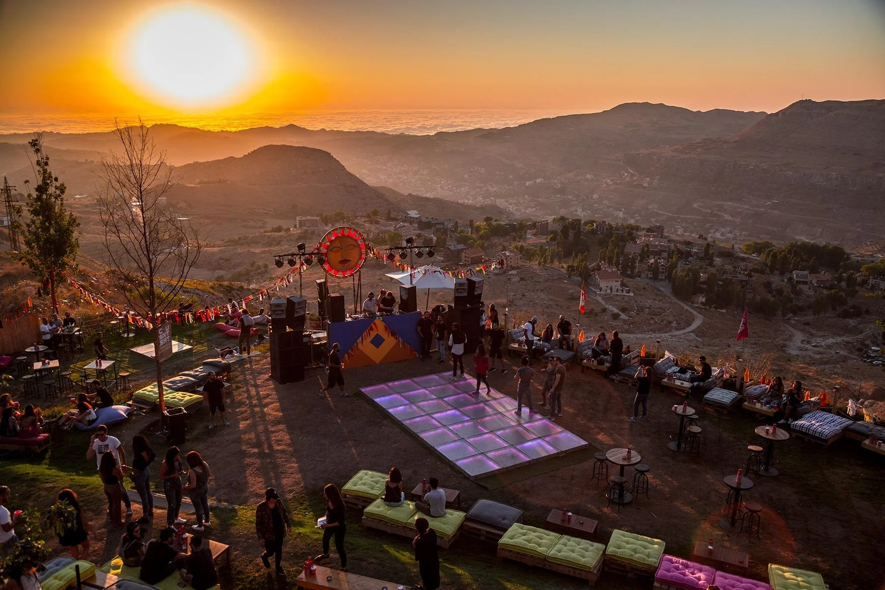 Sunset view over a hilly landscape with a party or event taking place on a terrace. There are people dancing on a light-up dance floor, sitting on couches, and socializing around tables. Decorated with flags and bunting, with a stage and DJ booth at the center.