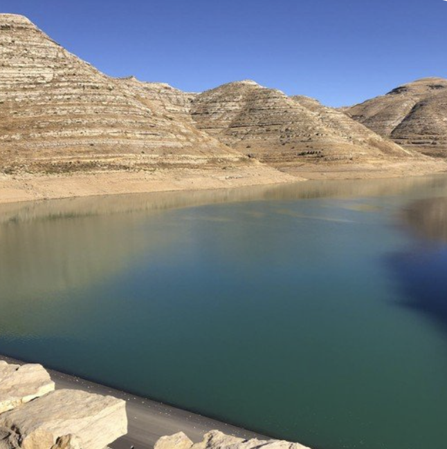 A calm body of water surrounded by arid, layered, beige and brown mountains under a clear blue sky.