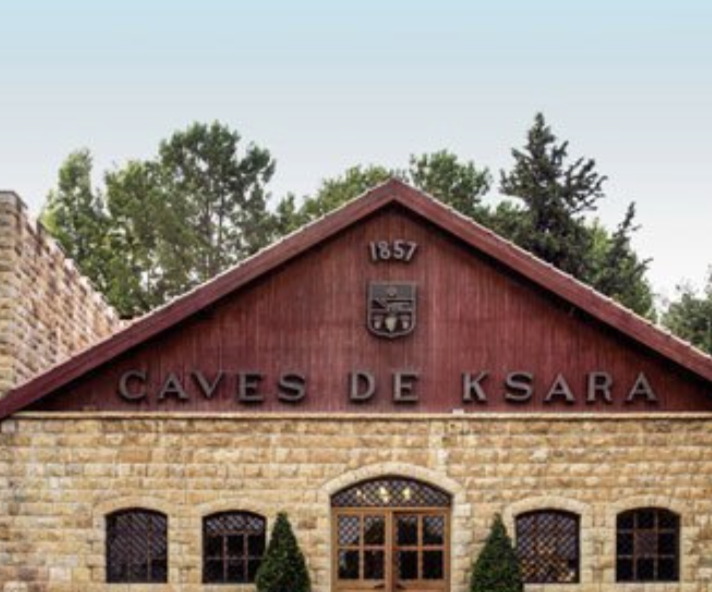 A stone building with a red wooden gable roof, displaying the words 'CAVES DE KSARA' and the year 1857, surrounded by trees.
