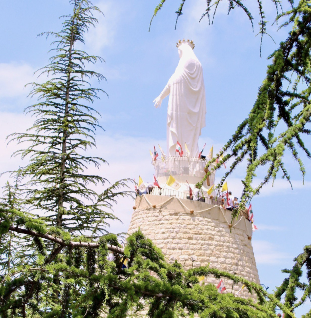 A statue of the Virgin Mary atop a stone pedestal, surrounded by trees and flags, with a blue sky background.