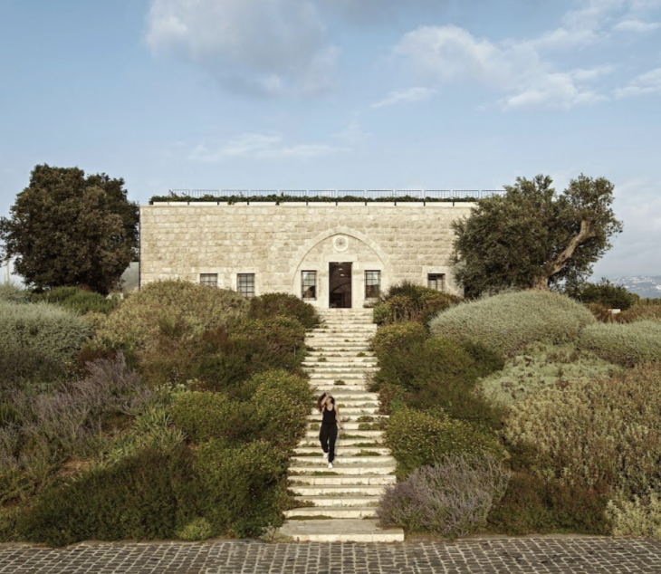 A stone staircase leading up to a historic stone building surrounded by greenery and bushes, with a woman walking down the stairs.