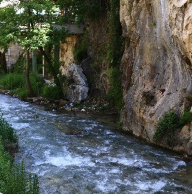 A narrow mountain stream flowing alongside a rocky cliff with some greenery, trees, and a bridge in the background.