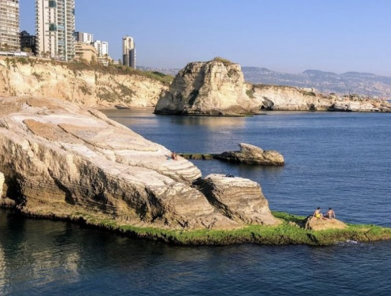 Rocky coastline with large rock formations and a small grassy area with people sitting, in a city near the water on a clear day.