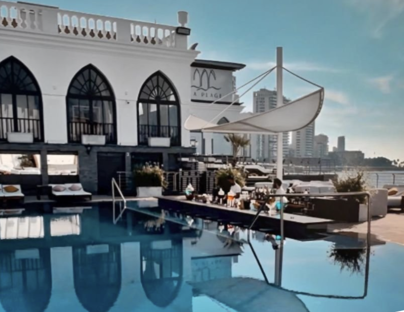 Luxury pool area at a waterfront restaurant with white building, lounge chairs, umbrellas, and city skyline in the background.