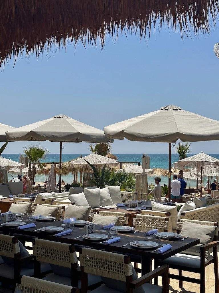 Beachside restaurant with tables set for dining, white umbrellas and lounge chairs, sandy beach, palm trees, and ocean in the background under a blue sky.