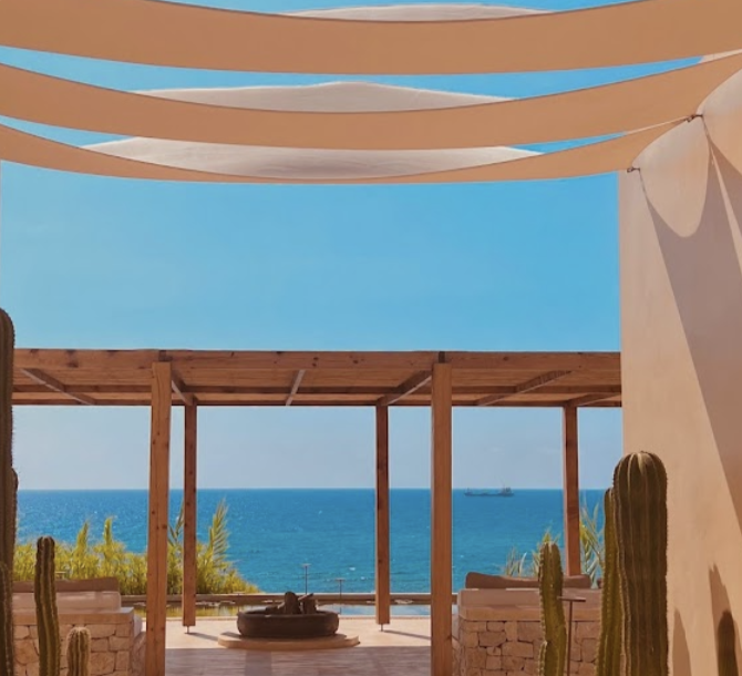 View of the ocean through a wooden pergola, with cacti and a shaded outdoor sitting area.