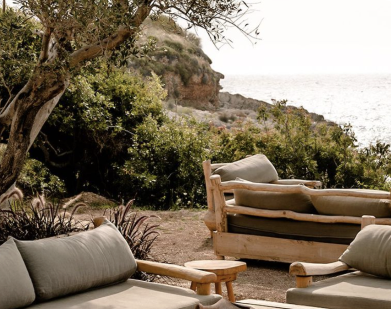 Coastal outdoor seating area with cushioned wooden furniture, trees, and a view of the ocean and rocky cliffs in the background